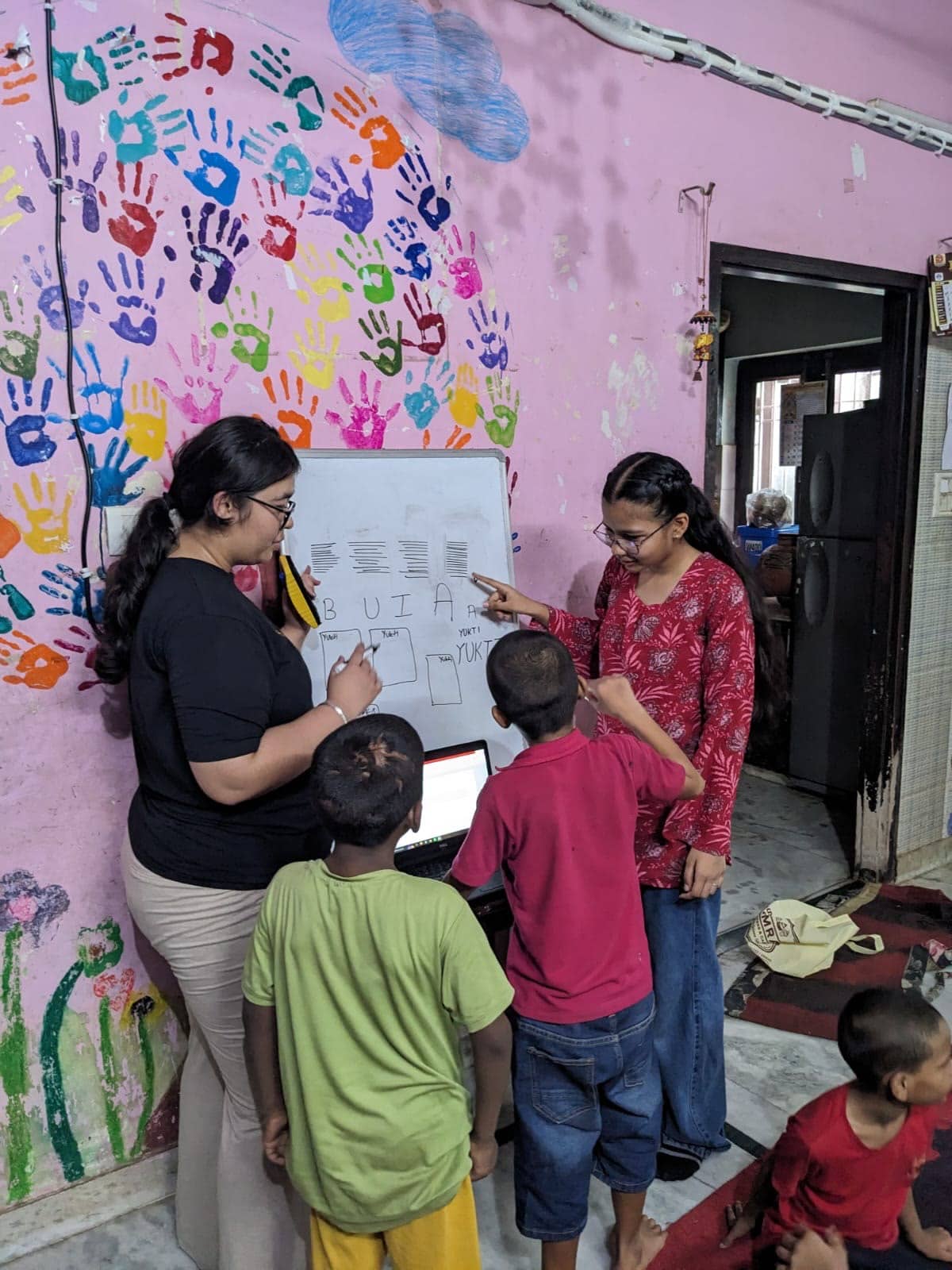 Volunteers at a whiteboard with children, handprints painted across the wall.