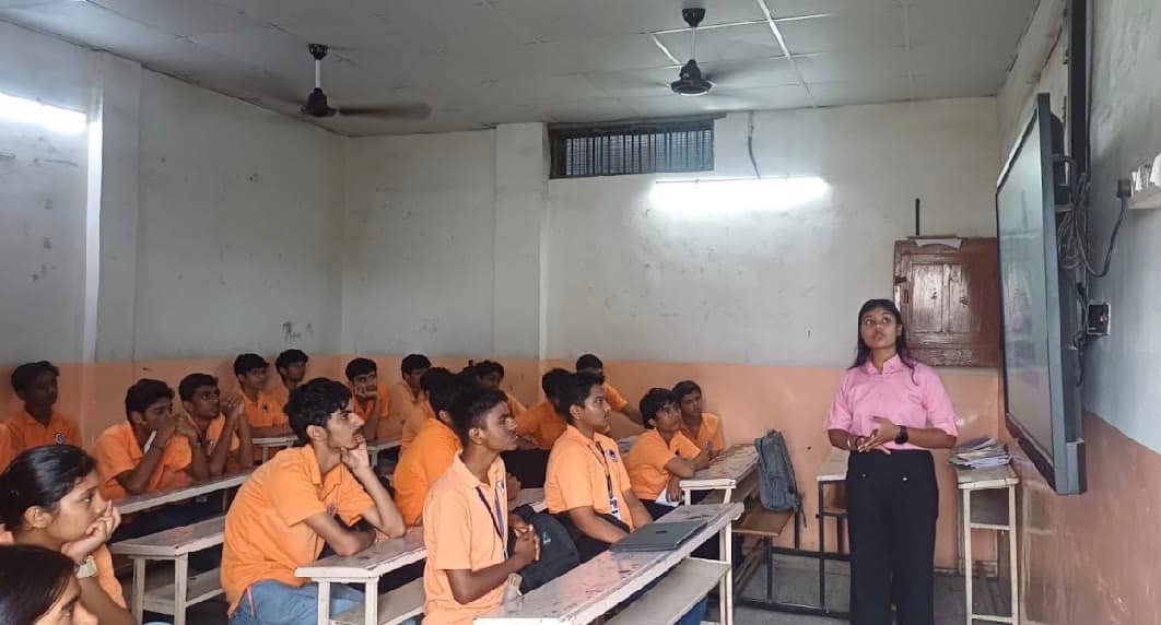 Students in orange uniforms in a classroom while a volunteer presents at the front.