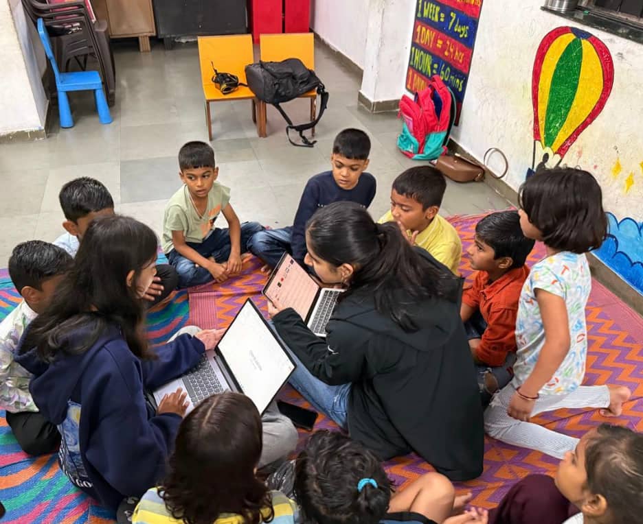 Overhead view of two volunteers on a patterned mat with about ten children, laptops open, a painted hot-air balloon on the wall.
