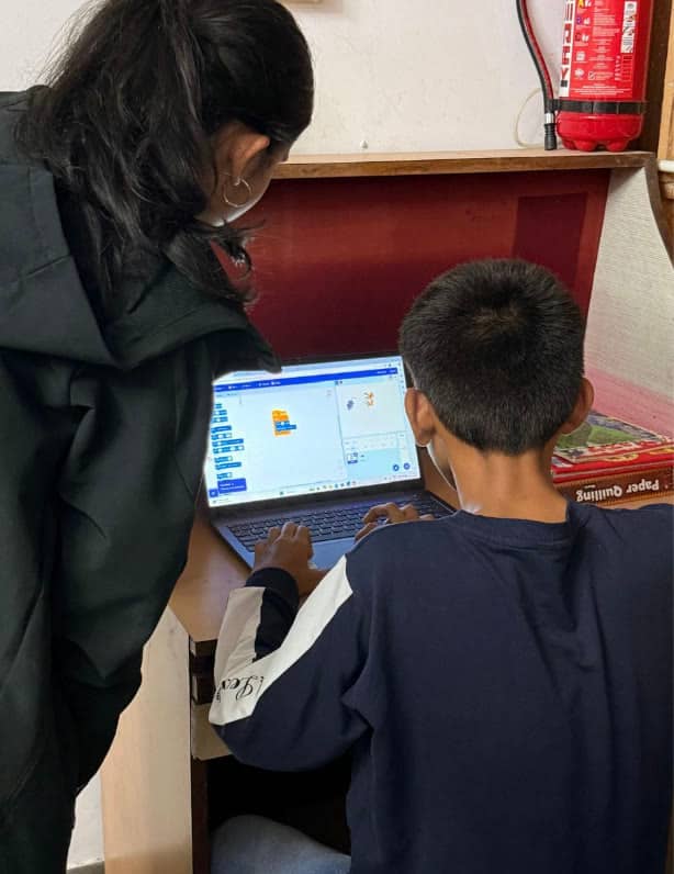 A volunteer leans over a boy at a laptop running Scratch, seen from behind.